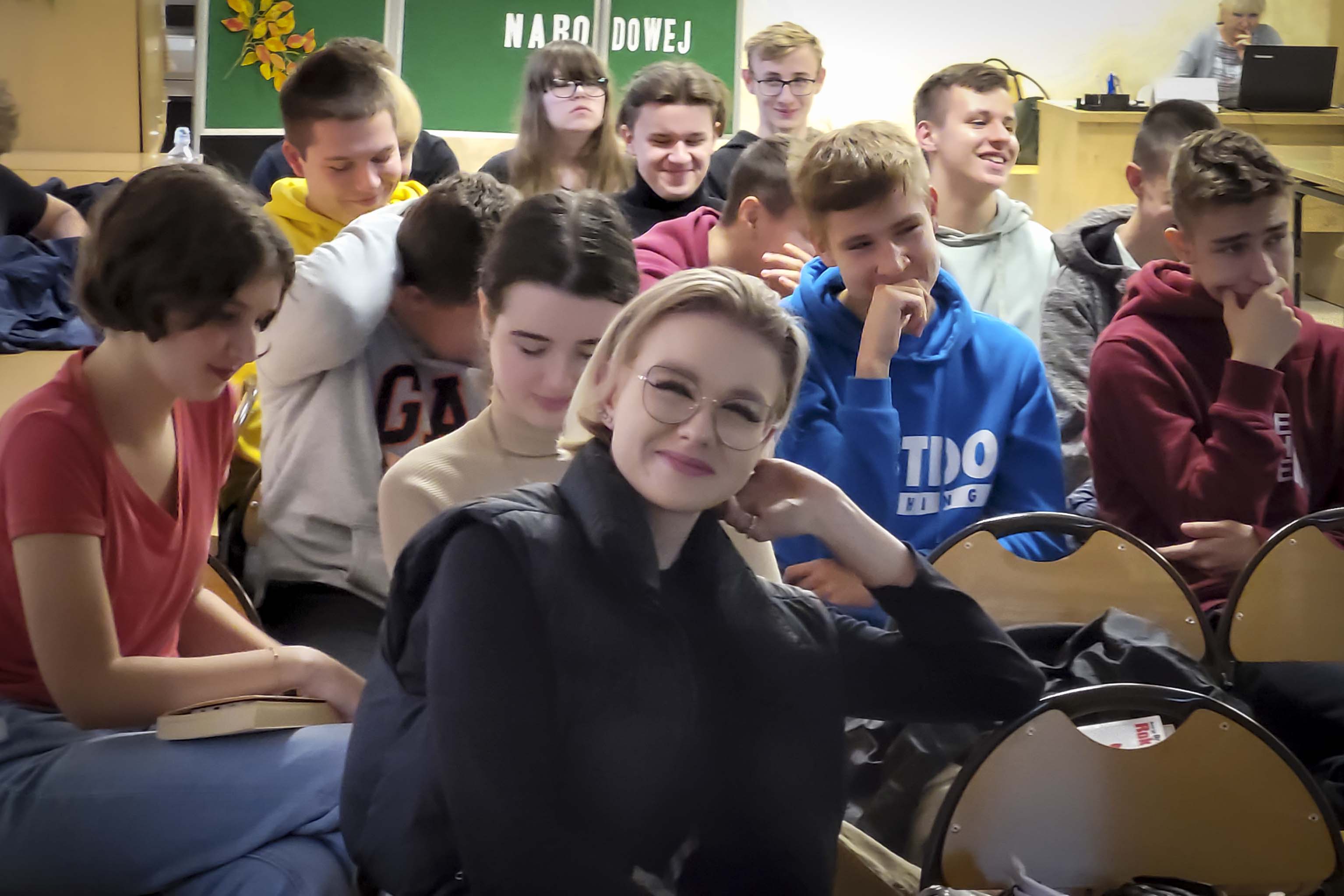 Uśmiechnięta dziewczyna w okularach siedzi na krześle patrzy w obiektyw w tle grupa dziewczyn i chłopców