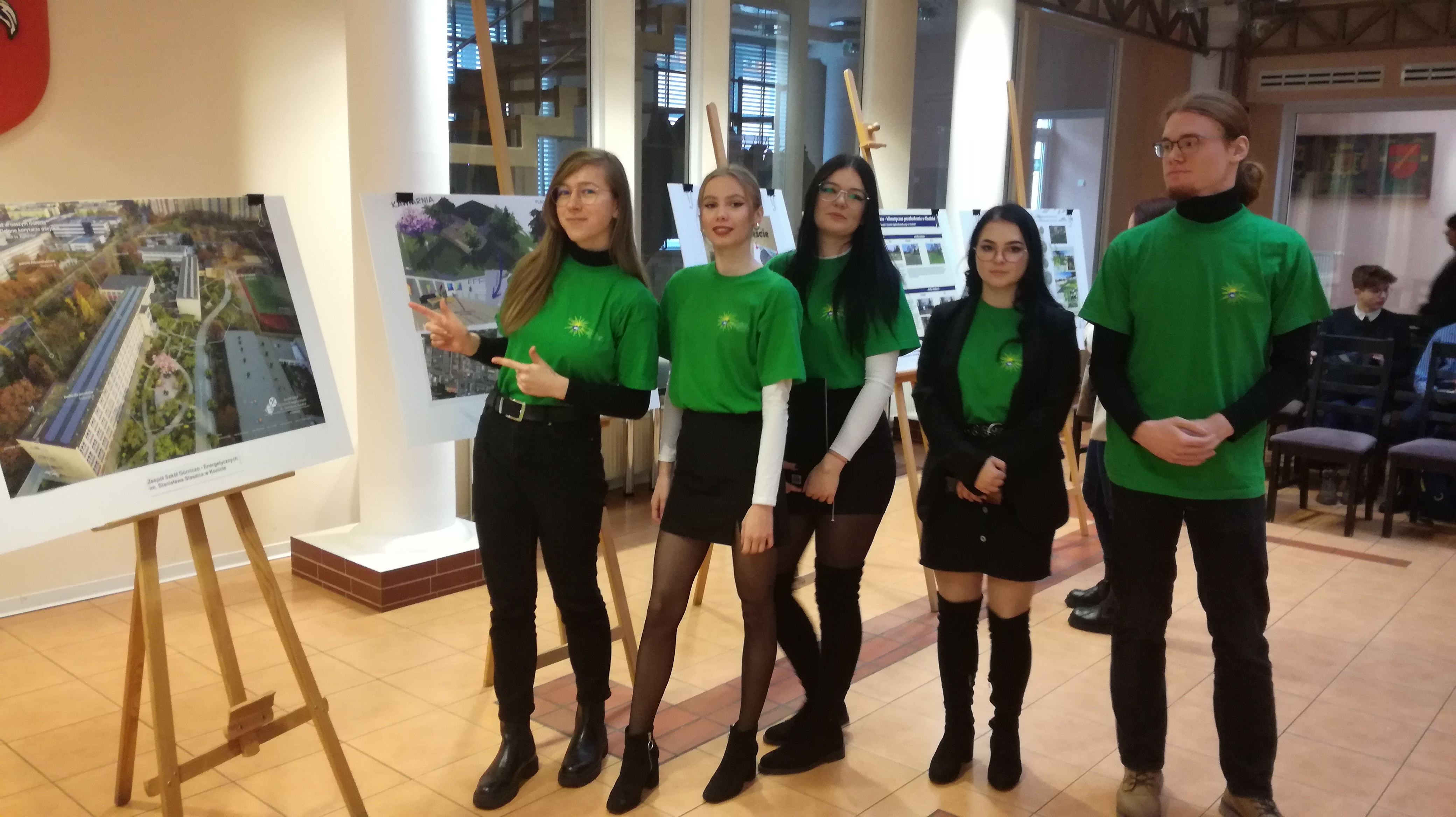 cztery dziewczyny i jeden chłopak w zielonych koszulkach, jedna dziewczyna wskazuje na ilustrację stojącą na sztaludze